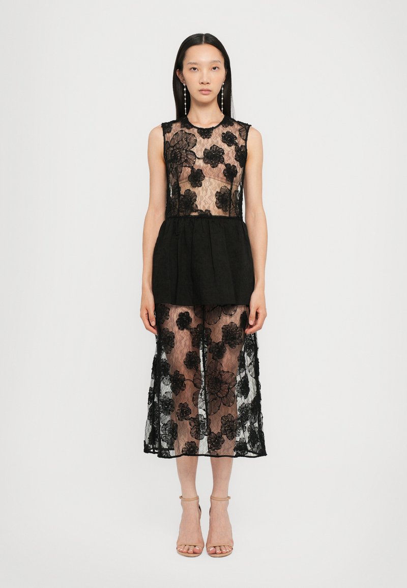 Vestido de encaje negro con patrones florales, con un cuerpo semi-transparente, cintura de peplum y una falda fluida. Combinado con sandalias de tacón nude.