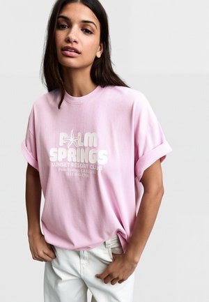 Mlada žena s dugom tamnom kosom nosi svijetloružičastu majicu s podvrnutim rukavima na kojima je ispisano "Palm Springs Sunset Resort Club" te bijele hlače, na jednoličnoj pozadini.