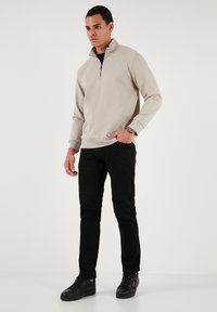 Svetlo béžový pulóver s dlhým rukávom a zipsom, kombinovaný s čiernymi nohavicami a tmavými teniskami. Hladký materiál s uvoľneným strihom.