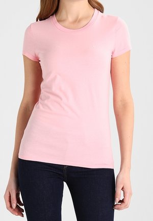 Žena oblečená v těsně padnoucím světle růžovém tričku s krátkým rukávem a tmavě modrých džínách stojí před jednoduchým světlým pozadím.