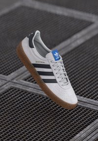 Grå ruskinds sneakers med sorte striber, struktureret gummisål og blåt logo-detalje. Har perforeringer og guld "MONTREAL" tekst på siden.