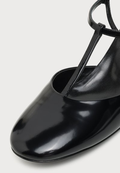 Chaussure en cuir noir brillant avec bride en T, bout arrondi et fine bride de cheville sur fond clair uni.
