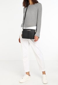 Svart läder crossbody-väska med dragkedjestängning, slät yta och subtila sömdetaljer. Bärs med vita byxor och grå tröja.