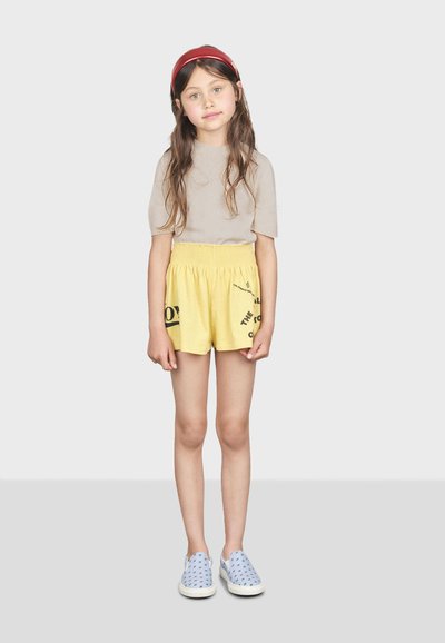 Pantalones cortos de algodón amarillos con cintura elástica y estampado gráfico negro, combinados con una blusa de manga corta en color beige claro y zapatos azul de tipo slip-on.