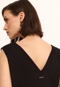 Czarna sukienka bez rękawów z dekoltem w szpic, gładka faktura i mała metka marki blisko tyłu. Złoty dodatek widoczny na uchu.