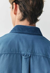 Blå bomulds skjorte med en kordfløjlskrave, der har stikninger over skuldrene og en stofsløjfedetalje på bagsiden.