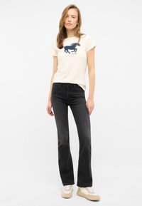 Camiseta crema con un gráfico de caballo en azul marino, combinada con jeans ajustados y desgastados en negro y zapatillas beige. Diseño simple y estilo casual.