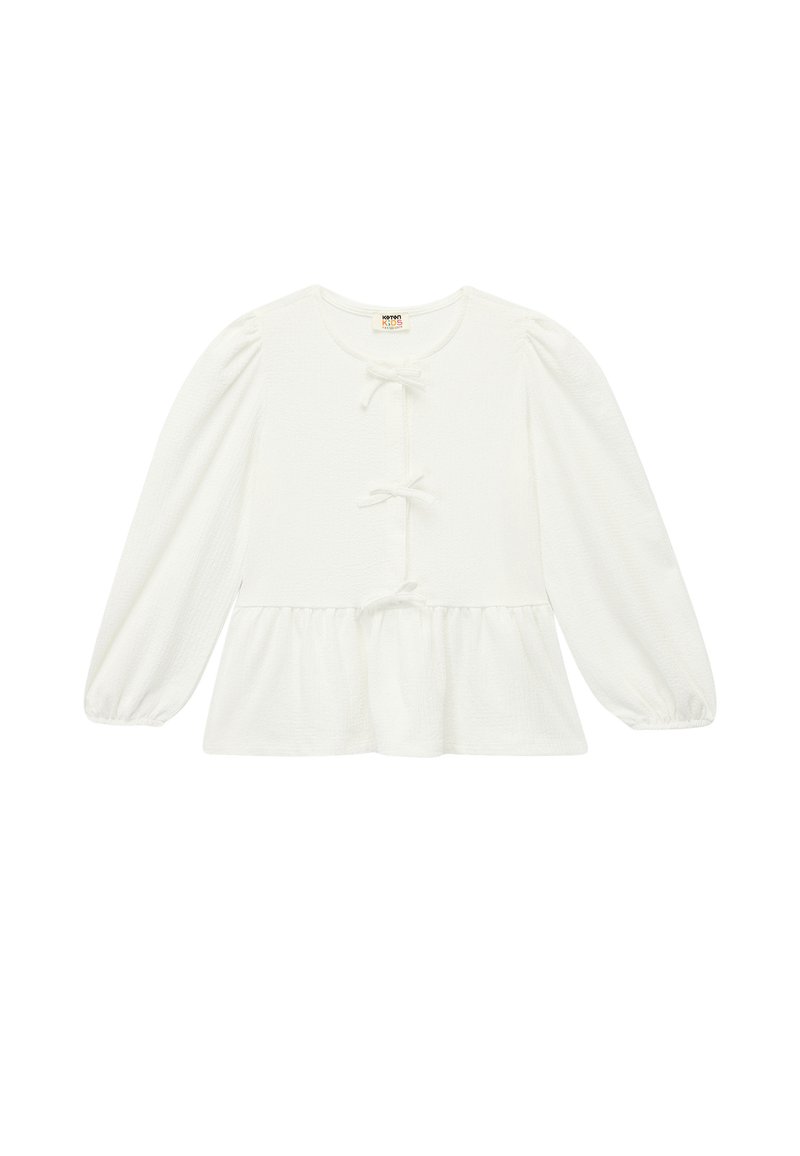 Haut à manches longues blanc en tissu texturé. Il présente un col rond, une fermeture à boutons avec des détails en nœud, et un ourlet peplum.