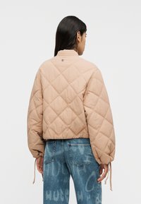 Μπεζ κοντό μπουφάν καπιτονέ με ψηλό γιακά και ελαστική μέση. Διαθέτει ρυθμιζόμενα κορδόνια στα μανίκια. Φορεμένο με μπλε τζιν.