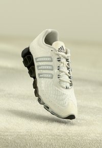 White Adidas running shoe with black segmented sole, mesh upper, and three signature stripes against soft beige textured background.