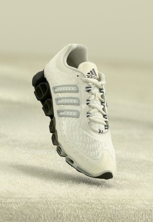 Zapatilla de running blanca de Adidas con suela segmentada negra, parte superior de malla y tres rayas distintivas sobre un fondo texturizado beige suave.