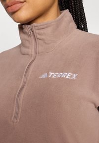 Sudadera de forro polar marrón claro con cuello alto y media cremallera. Características destacadas incluyen el logo bordado "TERREX" y una textura suave.