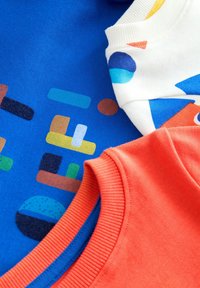 Tre magliette colorate esposte insieme: blu con motivi astratti, bianca con forme geometriche e arancione con un colletto a coste.