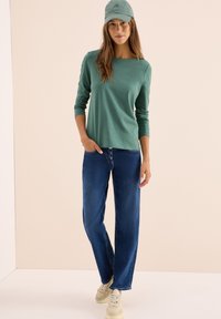 Kvinna klädd i grön långärmad skjorta, grön keps, blå jeans med knappgylf och beige sneakers, står med en hand i fickan.
