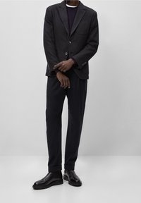 Uomo in blazer scuro, pantaloni neri e stivali di pelle nera in piedi contro uno sfondo grigio semplice.