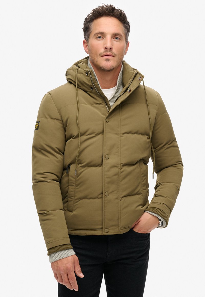 Chaqueta acolchada verde oliva con capucha, que cuenta con una cremallera en la parte frontal, botones de presión y puños acanalados. Textura suave con detalle del logo en la manga.