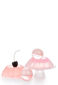 Trois contenants de crème rose semblables à de la gelée, avec des sommets arrondis, présentés sur des supports en verre transparent. Une cerise noire brillante décore la scène.