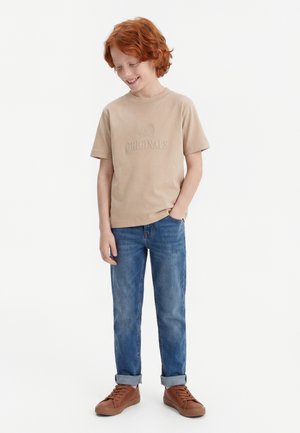 Garçon roux portant un t-shirt beige, un jean bleu avec des revers et des baskets marron, debout avec une main dans la poche et souriant vers le bas.