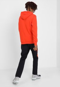Sudadera con capucha roja, pantalones negros con franjas naranjas, zapatillas gris. La tela parece suave; el diseño es casual y ajustado.