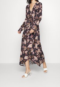 Žena v černých šatech s dlouhým rukávem a po kotníky, pokrytých růžovými a oranžovými paisley vzory, a bílých sandálech na podpatku s otevřenou špičkou.