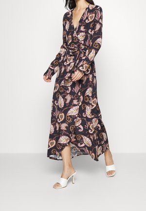 Vrouw in een zwart langslaven jurk met enkel lengte, versierd met roze en oranje paisley patronen, en witte open teen hakken.