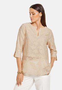 Beige Bluse mit V-Ausschnitt und Dreiviertelärmeln, die mit strukturierten Streifen und floralen Mustern verziert ist. Kombiniert mit weißen Hosen und einem goldenen Armband.