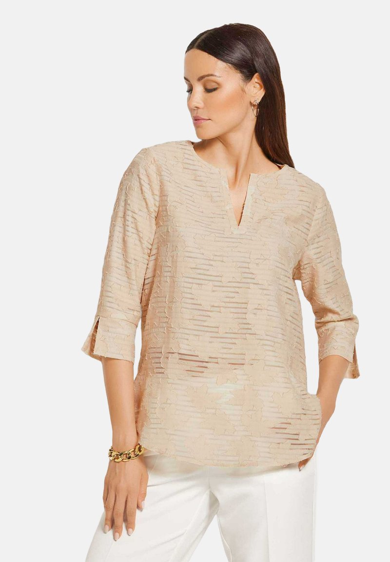 Beige Bluse mit V-Ausschnitt und Dreiviertelärmeln, die mit strukturierten Streifen und floralen Mustern verziert ist. Kombiniert mit weißen Hosen und einem goldenen Armband.