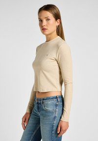 Beige, ribbet langærmet crop top med rund halsudskæring og subtilt logodetalje; parret med lyseblå, tætsiddende jeans.