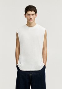 Weißes ärmelloses Baumwolloberteil mit rundem Halsausschnitt, rohem Saum und seitlichen Schlitzen, kombiniert mit dunkelblauen Jeans. Einfaches und minimalistisches Design.
