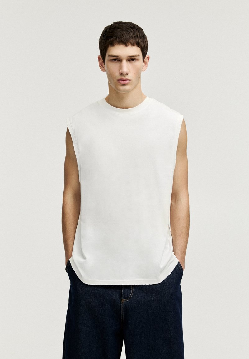 Weißes ärmelloses Baumwolloberteil mit rundem Halsausschnitt, rohem Saum und seitlichen Schlitzen, kombiniert mit dunkelblauen Jeans. Einfaches und minimalistisches Design.