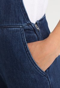 Mörkblå denimöveralls med siddragkedja och synlig sömnad. Texturen är slät, vilket framhäver plaggets hållbara design.