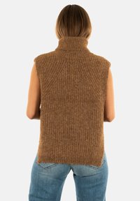 Femme aux cheveux bruns mi-longs portant un pull col roulé sans manches marron et un jean bleu clair, de dos.