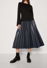 Haut noir à manches longues associé à une jupe plissée foncée en matériau brillant, complété par des chaussettes noires et des mocassins cirés.