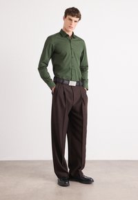 Cămașă închisă la culoare, cu guler, centură neagră cu cataramă metalică și pantaloni plisati maro. Pantofi negri completează ținuta.