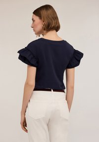 Maglietta blu scuro a maniche corte con maniche arricciate, abbinata a pantaloni chiari e una cintura marrone. Design semplice e casual.