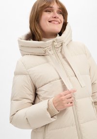 Beige pufferjack met een hoge kraag en oversized capuchon. Voorzien van een ritssluiting aan de voorkant, trekkoorden en ribgebreide mouwen. Glad textuur.