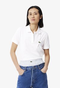 Polo shirt bianco in cotone con una patta a tre bottoni e logo verde firmato sul petto sinistro. Indossato con pantaloni corti in denim blu a vita alta.