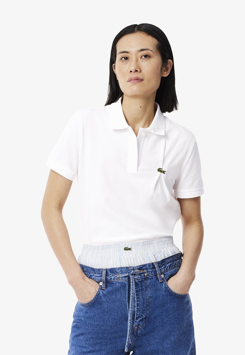 Witte poloshirt van katoen met een drie-knopssluiting en het kenmerkende groene logo op de linkerborst. Draagt een blauwe denim high-waisted short.