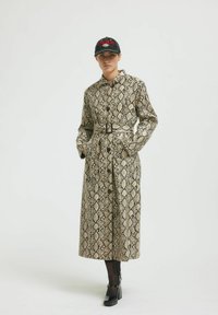 Plašč s krokodiljim vzorcem v bež in rjavih odtenkih, z pasom, gumbnim zapiranjem in dolgimi rokavi, v kombinaciji s črnim klobukom in čevlji z debelim petkom.