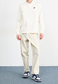 Krämfärgad långärmad polotröja med svart Nike-logotyp, kombinerad med ljusbeige cargobyxor och marinblå och vita sneakers. Texturen är mjuk och avslappnad.