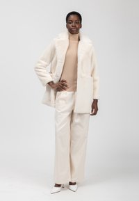 Cappotto in finto pelo color crema con ampio colletto, chiusura a bottoni e tasche anteriori, abbinato a un dolcevita beige e pantaloni a gamba larga di un beige chiaro.