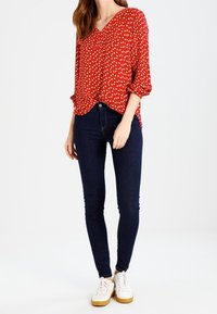 Blouse rouge avec des motifs en forme de cœur blancs, décolleté en V, manches retroussées ; associée à un jean skinny foncé et des sneakers blancs.