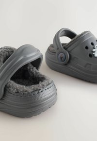 Chaussures de style sabot grises avec un doux revêtement en polaire grise, un extérieur en caoutchouc texturé et une sangle ajustable. Des trous de ventilation et un détail en bouton sont visibles.