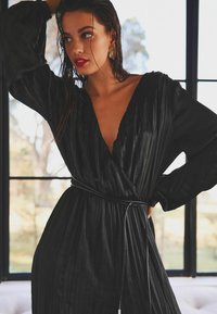 Schwarzes, plissiertes Kleid mit tiefem V-Ausschnitt und langen Ärmeln, tailliert mit einem dünnen schwarzen Gürtel. Glatte Textur und leichtes Material.