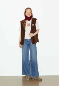 Gilet en polaire marron sur un T-shirt graphique blanc, associé à un jean bleu à jambe large et des bottes à imprimé léopard. Un bonnet en tricot rouge complète la tenue.