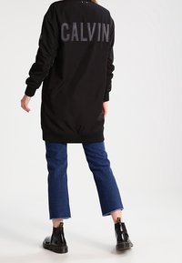 Czarna kurtka bombowiec z dużymi szarymi literami "CALVIN" na plecach, zestawiona z niebieskimi skrojonymi dżinsami i czarnymi błyszczącymi botkami za kostkę.