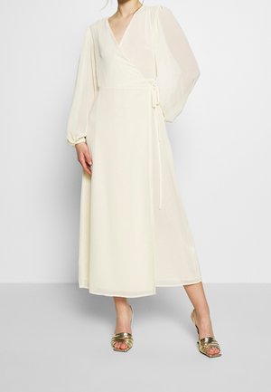 Frau in einem langen, cremefarbenen Wickelkleid mit transparenten langen Ärmeln und goldenen, offenen High-Heel-Sandalen, die vor einem schlichten Hintergrund steht.