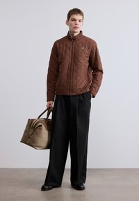 Καφέ μπουφάν με σχέδιο καπιτονέ, ψηλό γιακά, κλείσιμο με φερμουάρ και πλαϊνές τσέπες. Μαύρο παντελόνι φαρδύ στο κάτω μέρος και μπεζ τσάντα duffel με καφέ λεπτομέρειες.