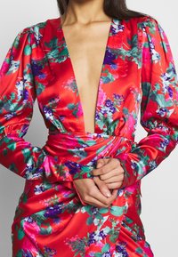 Robe florale en satin rouge avec décolleté en V profond, manches bouffantes et taille froncée. Des motifs vifs de rose, violet et vert ornent le tissu.