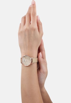 Person trägt eine beigen Stoffarmbanduhr mit einem hellen Holztextur-Zifferblatt und hält sanft sein Handgelenk mit der anderen Hand.